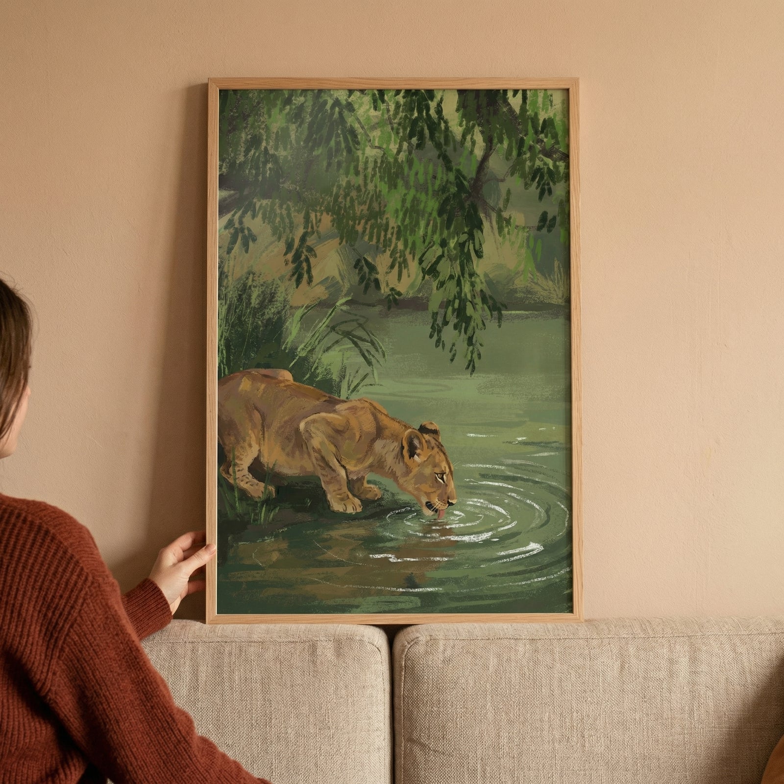 A vertical framed illustration held against a neutral wall depicts a young lion drinking from a serene body of water. The animal is rendered in warm tan and gold tones, leaning down from a grassy bank to reach the water's surface, which features delicate white ripples. Above, thick green foliage and overhanging leaves create a lush, shaded environment, with varying shades of green reflecting in the water below. The artwork is characterized by textured, expressive strokes that give the scene a tactile qualit