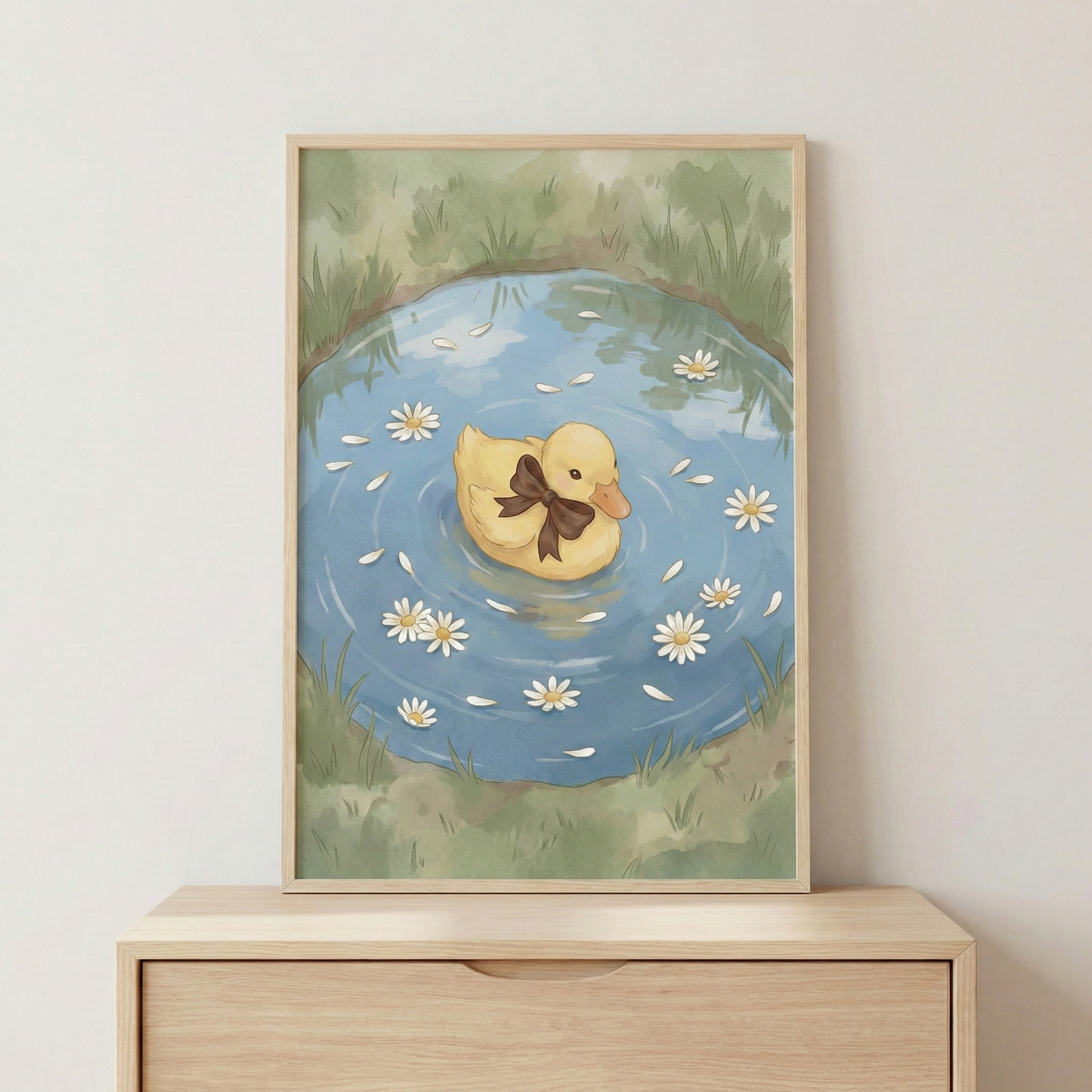 A framed illustration shows a small yellow duckling floating in the center of a round blue pond. The duckling wears a simple brown ribbon bow around its neck. White daisy flowers and individual white petals drift on the water's surface surrounding the duckling. The pond is bordered by a fringe of green grass. The artwork is presented in a light wood frame, which sits on a matching wooden dresser against a plain, light-colored wall. The style of the art is soft and whimsical.