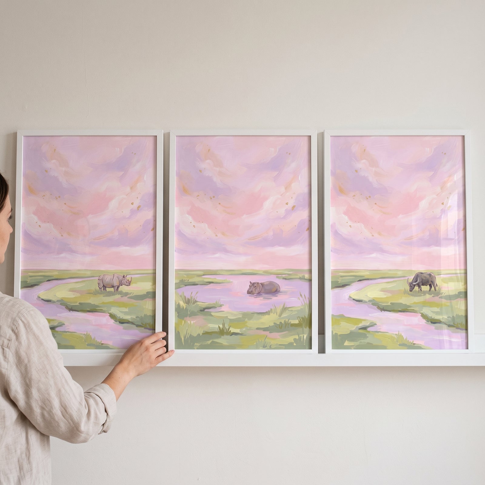 Hand adjusts a dusk sky safari animals wall art set of 3 with a rhino, hippo, and buffalo in pastel landscapes.