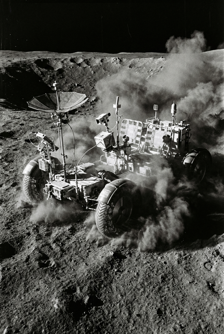 Lunar rover drives across the moon’s surface kicking up clouds of dust around its wheels, with antennas and equipment visible on top. The image captures motion and the rough, powdery texture of the terrain.
