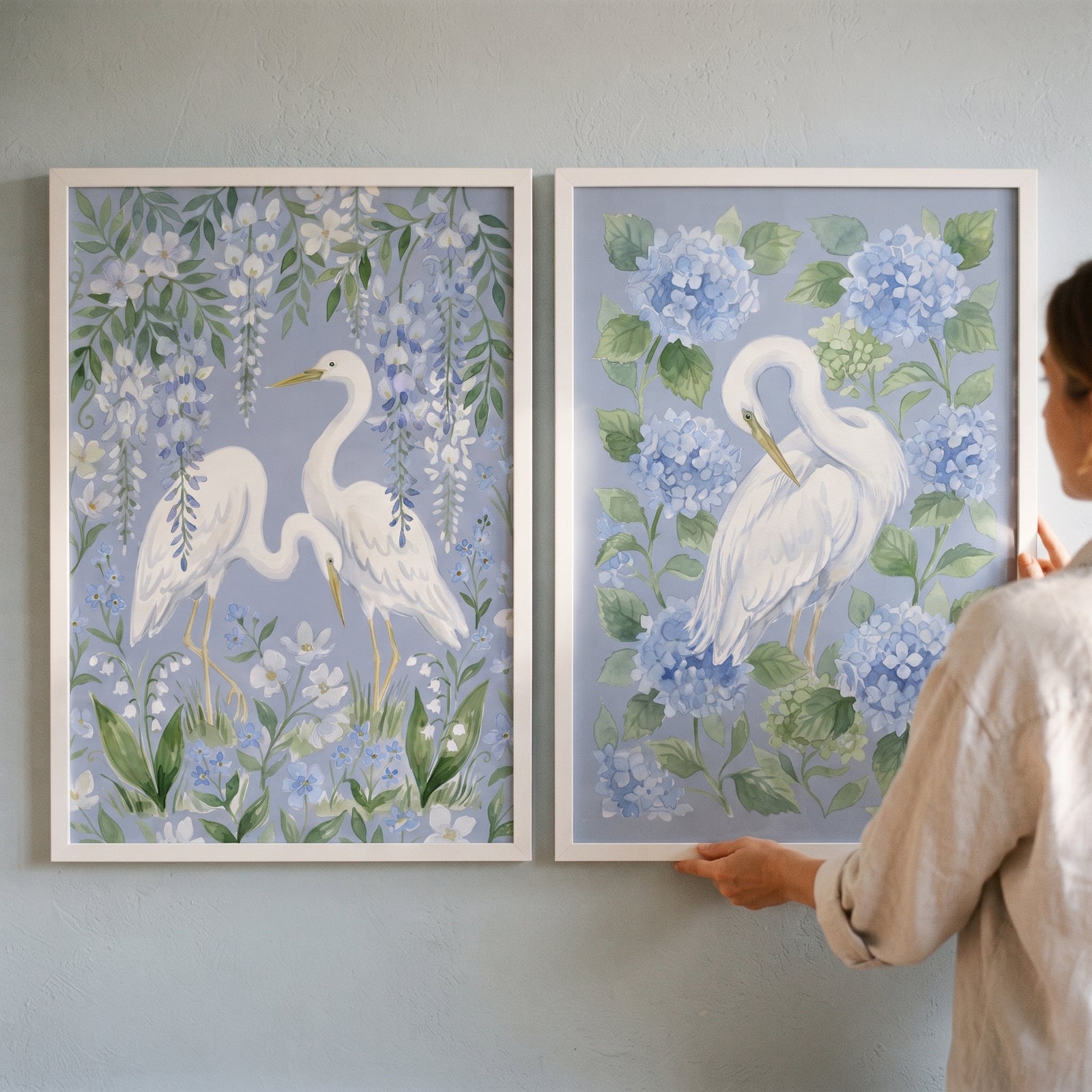 A person holds a dusty blue floral egret bird wall art set of 2 featuring white herons, hydrangeas, and wisteria.