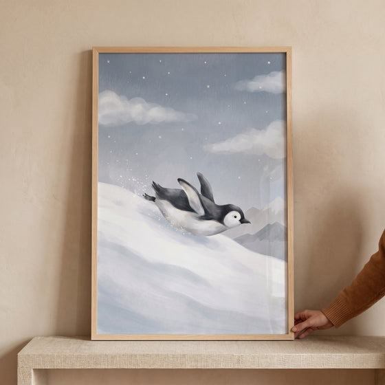 Dusty blue starry penguin wall art depicts a penguin sliding down a snowy slope in a wooden frame under a starry sky.