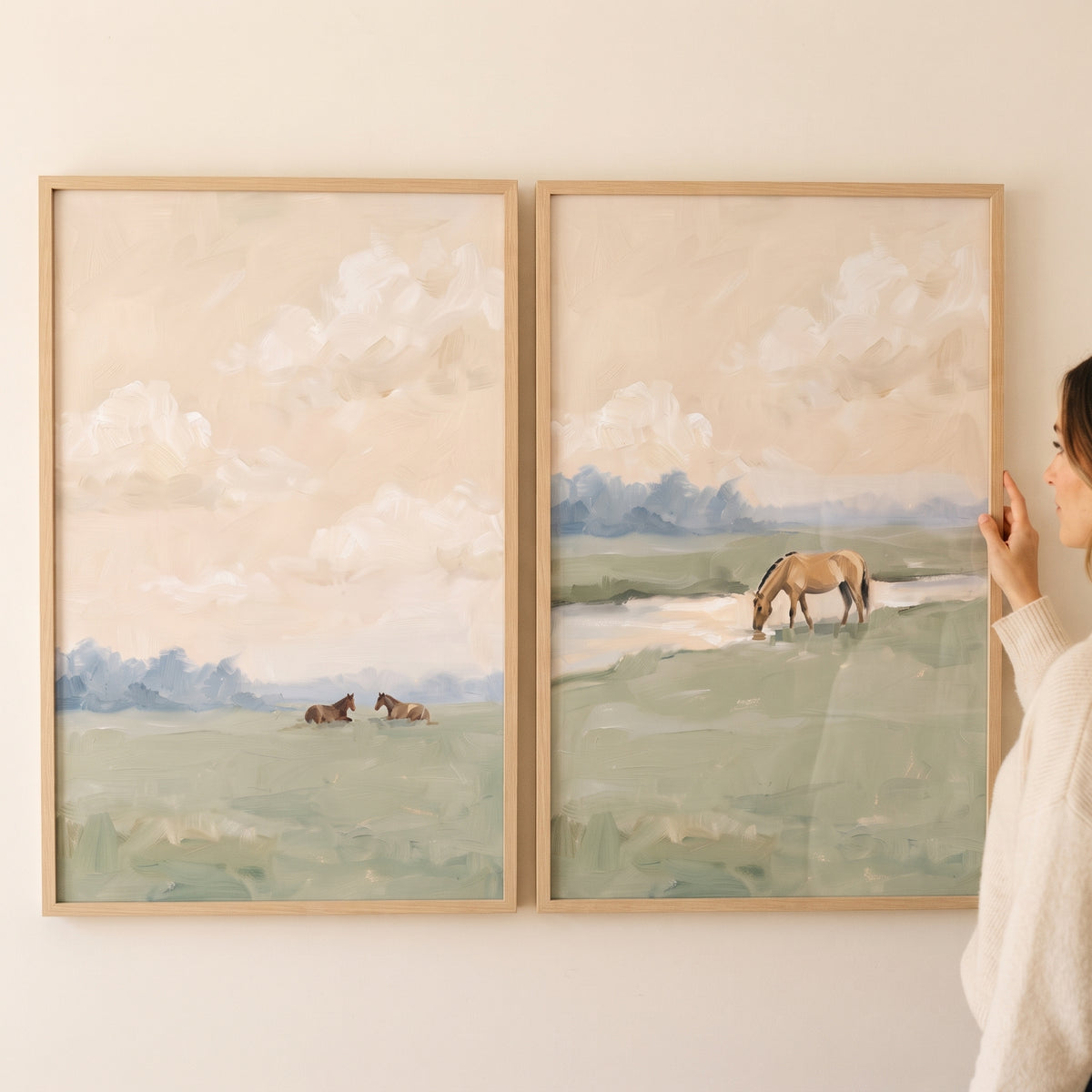 Hand adjusts a dusty green pastoral horse wall art set of 2 with horses in soft meadows under a cloudy beige sky.