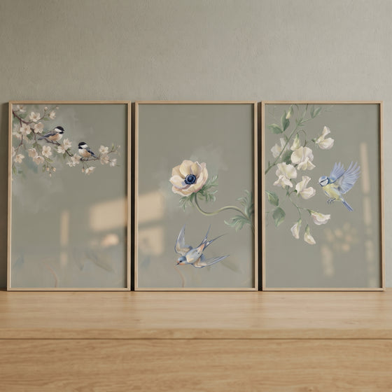 Dusty sage botanical bird wall art set of 3 with songbirds and white flowers in wood frames on a wooden surface.