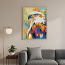 Modern living room with a gray sofa. Above the sofa hangs a framed, abstract, colorful painting of an eagle in profile, with white head feathers and vibrant orange, blue, purple body feathers. A brass floor lamp and a small plant on a side table are to the left.