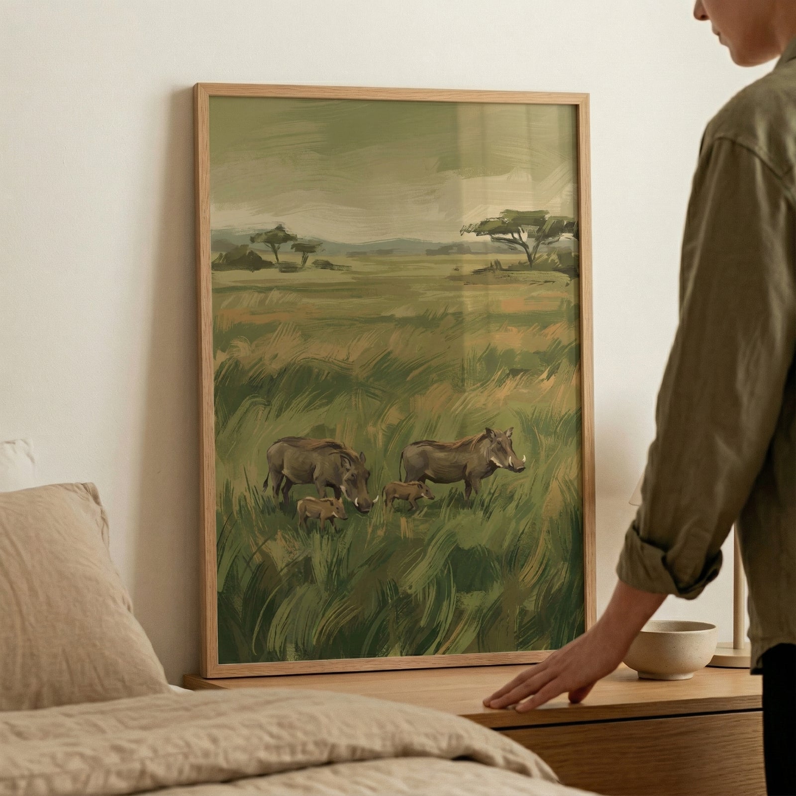 A vertical, light-colored wood frame containing an illustration of an African savanna landscape leans against a plain, neutral-toned wall. A person’s hand and arm are visible on the right, resting against the side of the frame. 
