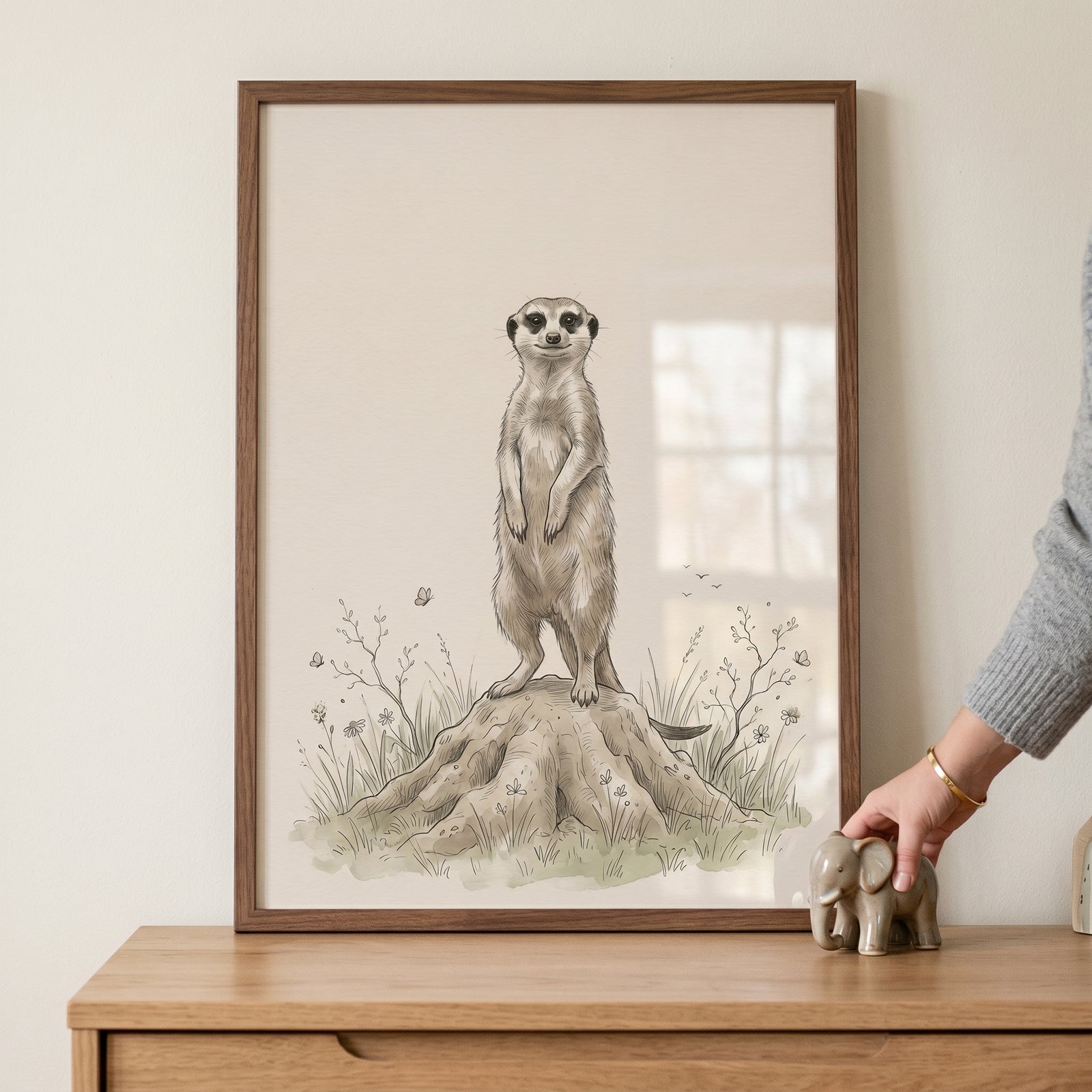 Framed earth tone standing meerkat wall art is on a wooden dresser beside a small elephant figurine and a person's hand.