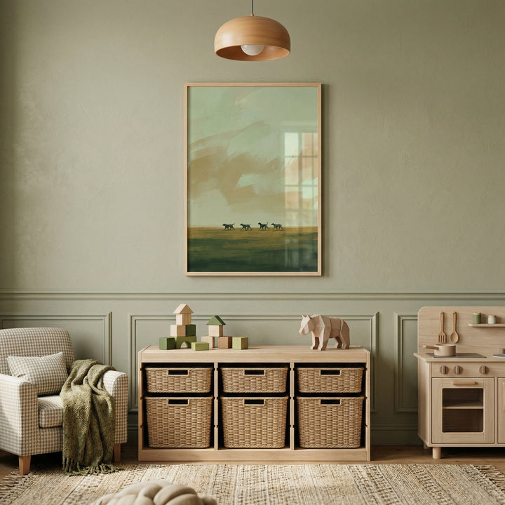 A beautifully designed, minimalist child's playroom with a serene sage green color scheme. The wall features elegant sage green wood paneling. Centered on the wall is a large wooden-framed artwork depicting an atmospheric abstract landscape. The illustration shows a hazy, sage and tan sky above a dark green field where four small animal silhouettes, like dogs, are walking in a line. 
Beneath the artwork is a low, light-wood shelving unit containing six woven wicker baskets.