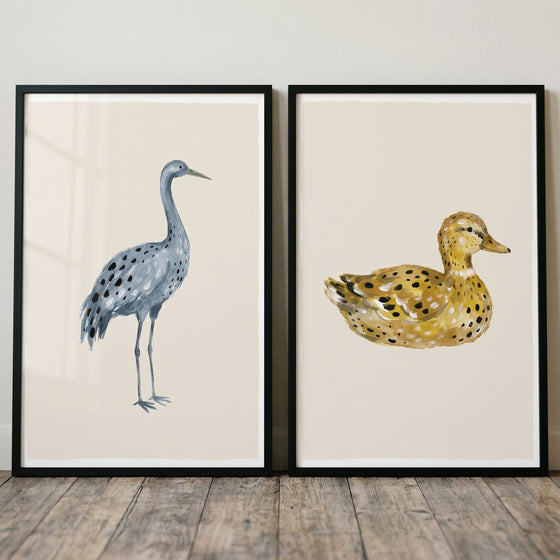 Earthy neutral stippled birds wall art set of 2 featuring a blue crane and a yellow duck in black frames on a wood floor.