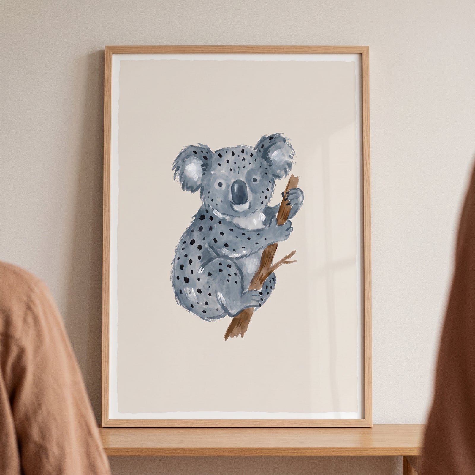 Earthy neutral watercolor koala wall art in a light wood frame shows a blue-grey koala holding a brown branch.