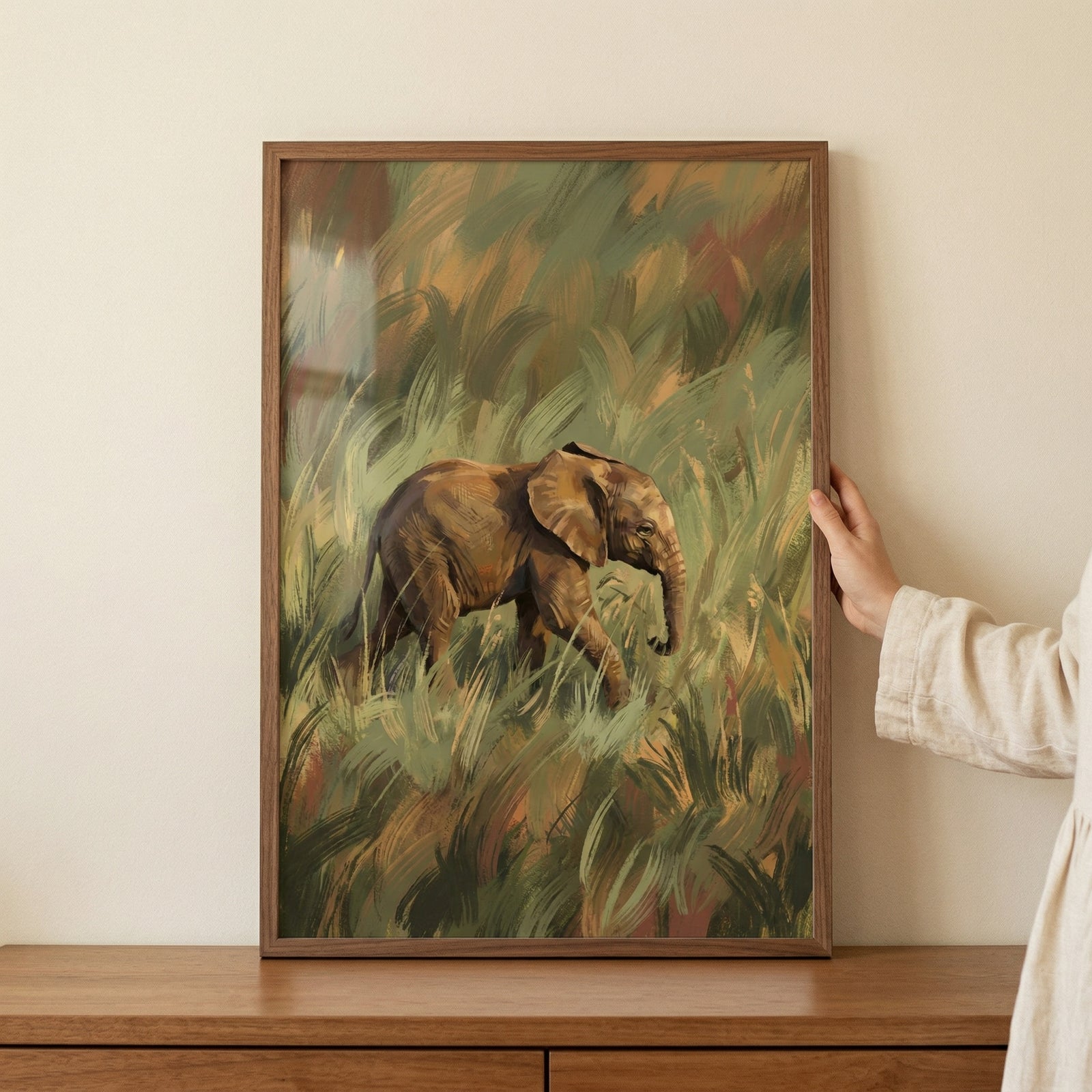 A framed vertical artwork depicts a small elephant walking through a field of tall, dense grass. The piece is rendered with expressive, visible brushwork in a palette of earthy tones, including shades of olive green, warm brown, and golden tan. 
