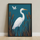 A framed graphic illustration features two white egrets in a stylized marsh setting. In the center, a large egret stands with a long, graceful neck and a pointed orange beak. It is surrounded by tall, brown marsh grasses and several cattails. In the upper left corner, a second, smaller egret is captured in mid-flight. The entire scene is set against a solid, deep teal background. The artwork is held within a simple black frame, which leans against a light-colored wall on a pale wooden floor.