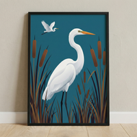 A framed graphic illustration features two white egrets in a stylized marsh setting. In the center, a large egret stands with a long, graceful neck and a pointed orange beak. It is surrounded by tall, brown marsh grasses and several cattails. In the upper left corner, a second, smaller egret is captured in mid-flight. The entire scene is set against a solid, deep teal background. The artwork is held within a simple black frame, which leans against a light-colored wall on a pale wooden floor.
