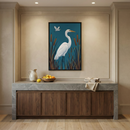 A sophisticated interior scene features a large, framed artwork of a white egret standing in a marsh against a teal background. The art is centered on a beige wall within decorative molding. Below the artwork is a dark wood cabinet with a thick grey marble countertop. On the counter sit a cream ceramic pitcher, a wooden bowl of citrus fruits, and folded linen napkins. The room has light wood floors and is illuminated by warm recessed ceiling lights.