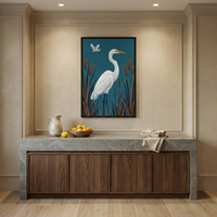 A sophisticated interior scene features a large, framed artwork of a white egret standing in a marsh against a teal background. The art is centered on a beige wall within decorative molding. Below the artwork is a dark wood cabinet with a thick grey marble countertop. On the counter sit a cream ceramic pitcher, a wooden bowl of citrus fruits, and folded linen napkins. The room has light wood floors and is illuminated by warm recessed ceiling lights.