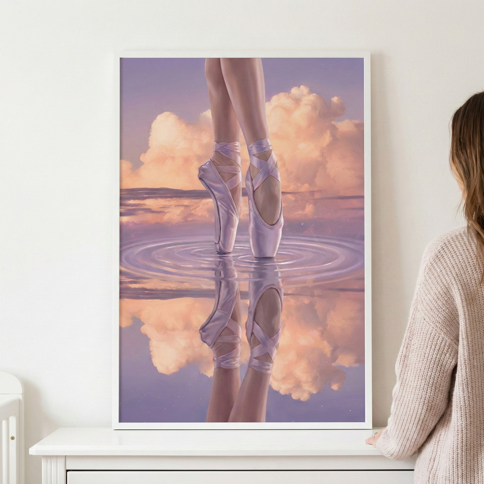 A framed artwork depicts a pair of slender legs in light pink satin pointe shoes, elegantly balanced on tiptoe upon a reflective water surface. The background is a dreamy, atmospheric sky with soft, billowing clouds in shades of rose, gold, and lavender. Below, the scene is mirrored in the water, creating delicate concentric ripples. The piece is held in a minimalist white frame on a plain wall, viewed by a person wearing a textured, light-colored sweater.