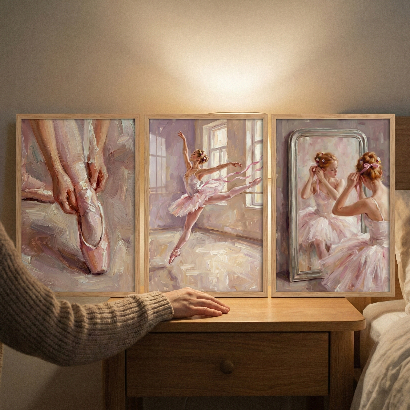 Three framed artworks are displayed side-by-side on a light-colored wooden nightstand, each capturing a moment from the world of classical dance. The pieces are created in a soft, impressionistic style with visible brushwork and a delicate palette of pinks, creams, and purples.The frame on the left shows a close-up depiction of hands carefully securing the satin ribbons of a ballet shoe. 