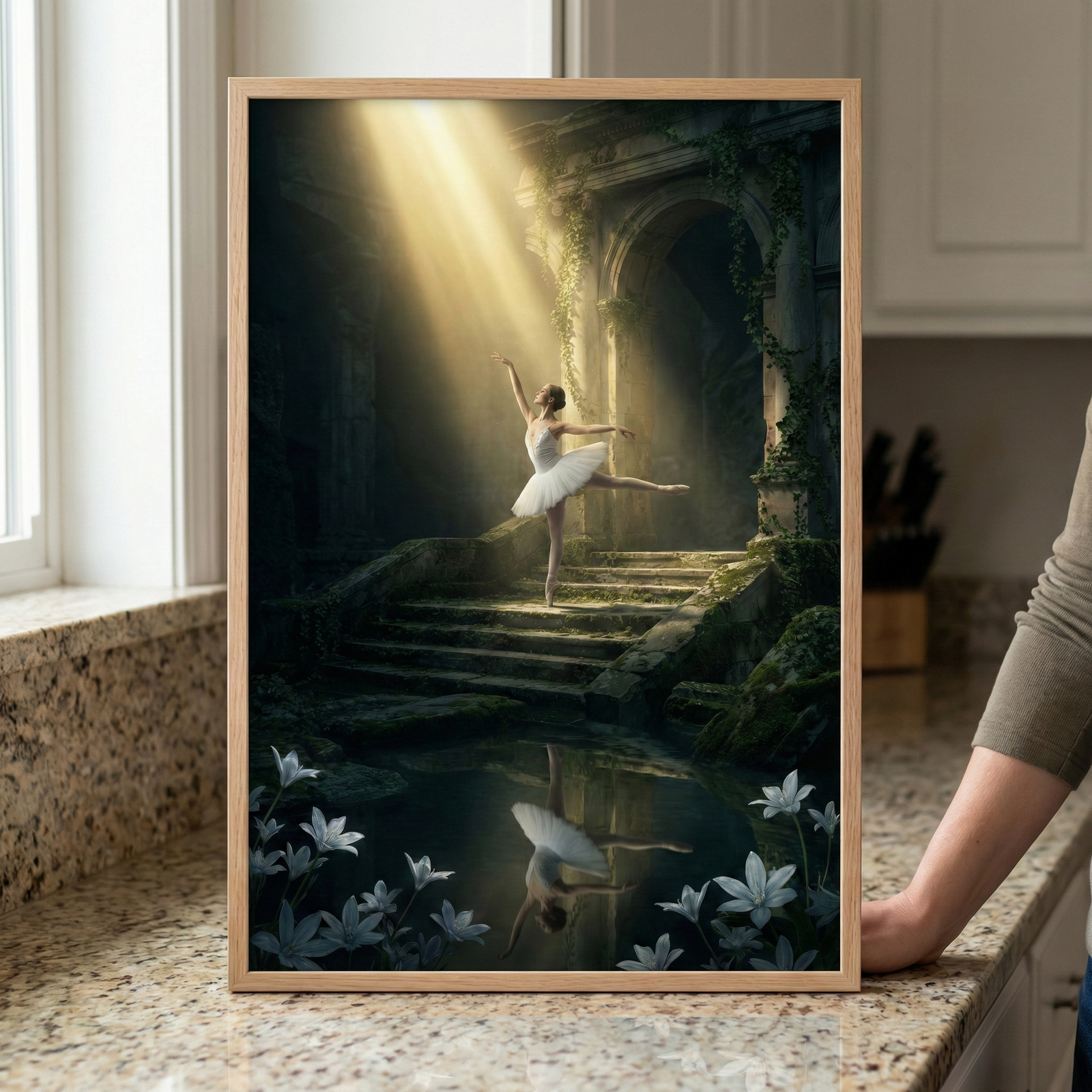 A framed vertical image depicts a ballerina in a white tutu striking a pose on moss-covered stone steps. She is illuminated by a dramatic shaft of golden light pouring from above within a vine-draped ancient stone ruin. Her reflection is visible in a dark pool of water in the foreground, surrounded by delicate white flowers. The artwork is held in a light wood frame and displayed on a granite surface indoors.