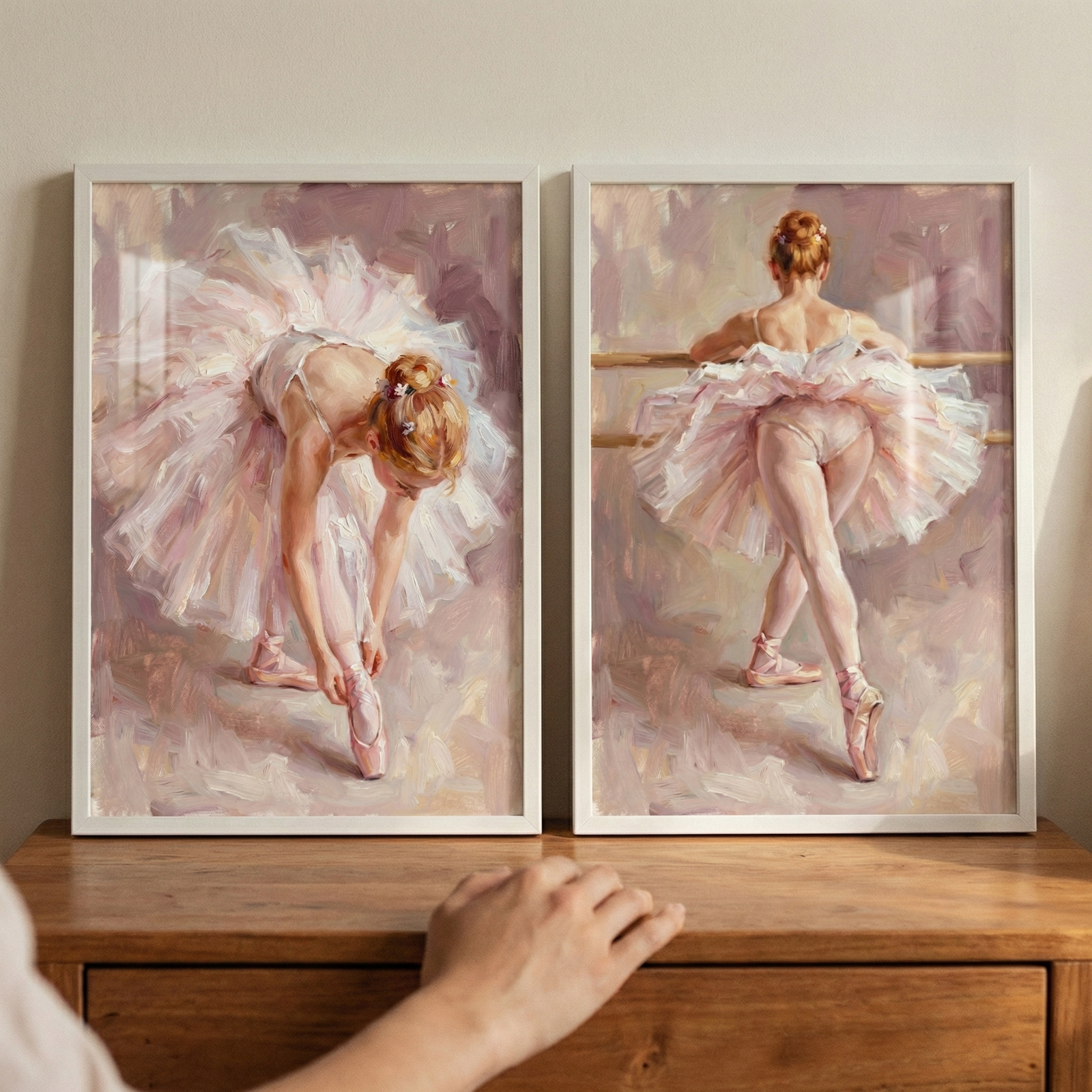 Two framed artworks in simple white frames rest side-by-side on a wooden dresser against a light wall. The piece on the left shows a young ballerina in a voluminous, ruffled tutu of soft pink and white, leaning forward to adjust the ribbons of her ballet shoe. The piece on the right captures a ballerina from behind, standing at a ballet barre with one leg gracefully extended.