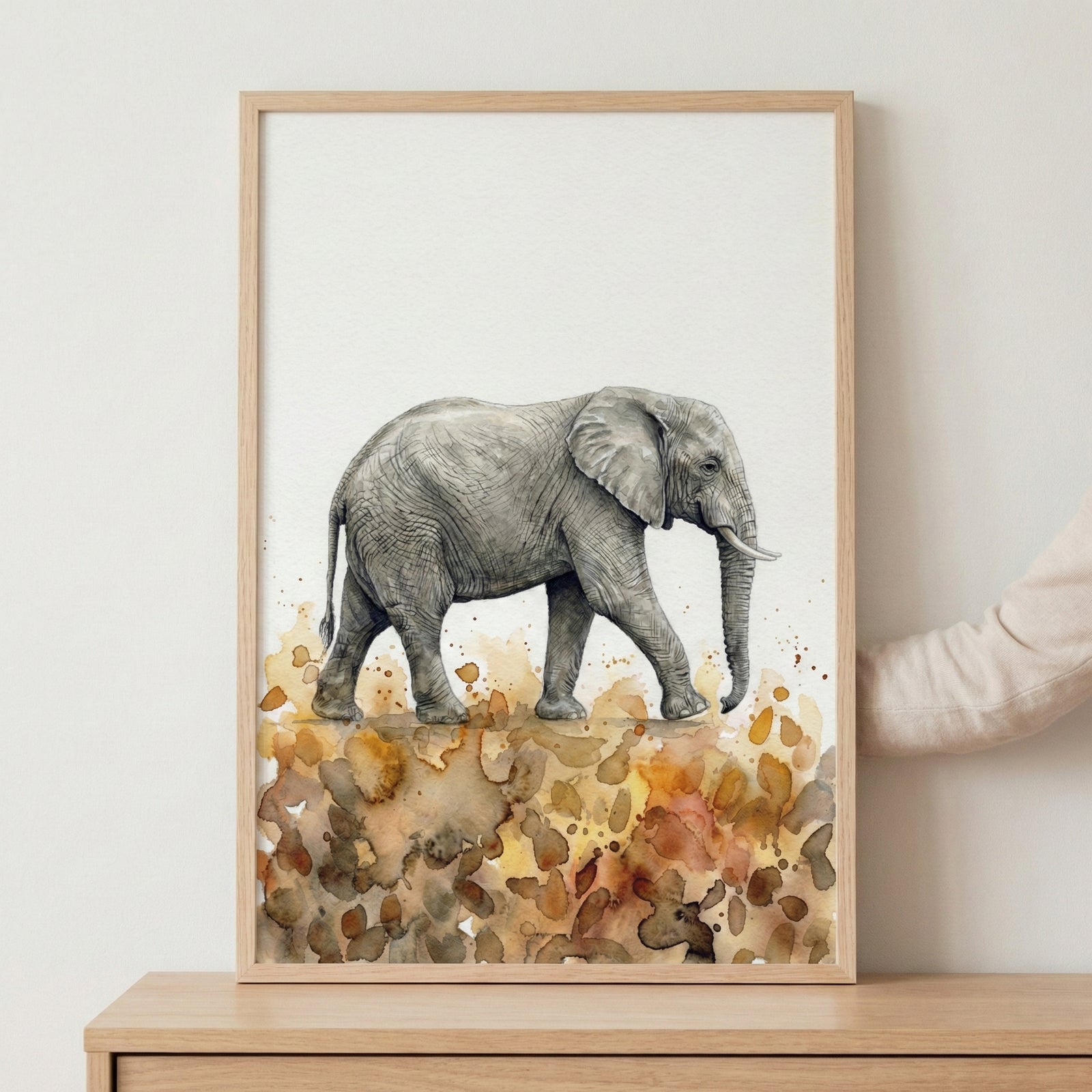 A person's hand holding a large framed watercolor painting of a giraffe against a neutral-toned wall.