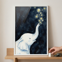 A framed watercolor-style illustration of a young white elephant is displayed on a light wooden shelf. The elephant is depicted from the side, with its trunk raised toward a dark, star-filled night sky. It appears to be blowing out glowing yellow stars and soft bubbles that float upward. The background transitions from a deep navy blue to a slightly lighter shade, textured with tiny white flecks representing distant stars. The artwork is housed in a simple white frame. 