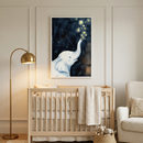 A serene, neutrally-toned nursery features a light wood crib with a cream knit blanket draped over the side. Centered above the crib is a large framed piece of wall art depicting a white watercolor-style elephant blowing glowing stars into a dark blue starry sky. A sleek, gold-finished arched floor lamp stands to the left. To the right, a textured cream armchair sits next to a small wicker basket containing two plush animals. The walls are finished with classic rectangular molding.