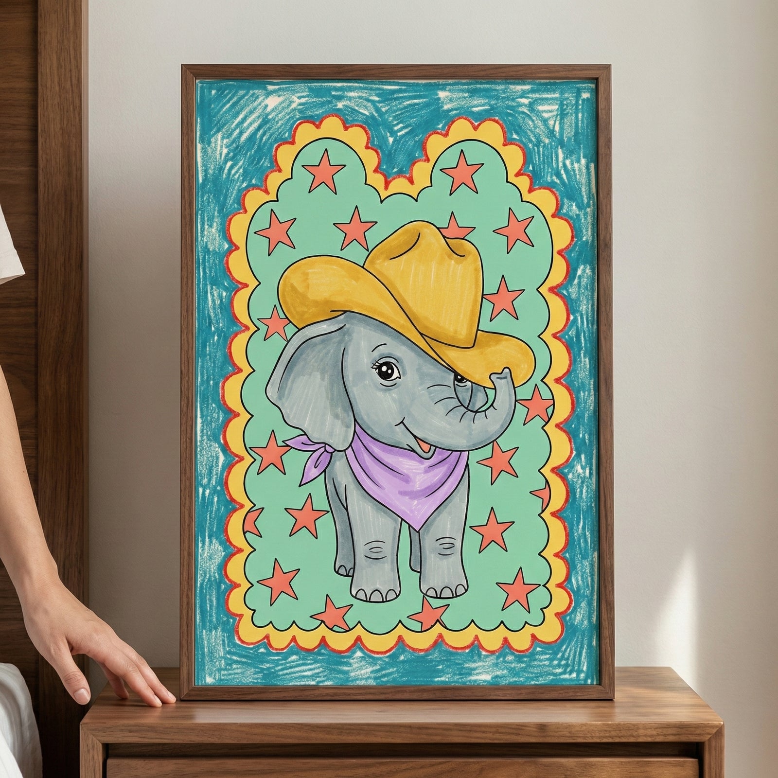 Framed drawing of a smiling cartoon elephant wearing a yellow cowboy hat and purple bandana, surrounded by red stars and a scalloped yellow and red border on a teal background, held by a person next to a wooden bed frame.