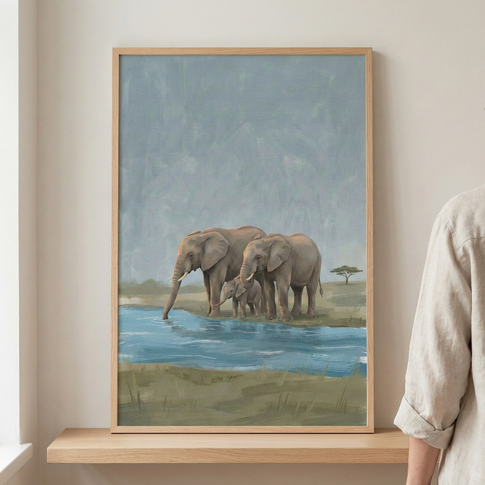 A framed piece of artwork rests on a light wood shelf. The illustration depicts a family of three elephants—two adults and a small calf—standing by a body of blue water. The scene is set on a grassy savanna under a muted, textured sky, with a single lone tree in the far distance. The frame is made of light wood, and the art has a soft, brushed texture. A person wearing a light-colored linen shirt is partially visible on the right side of the frame.