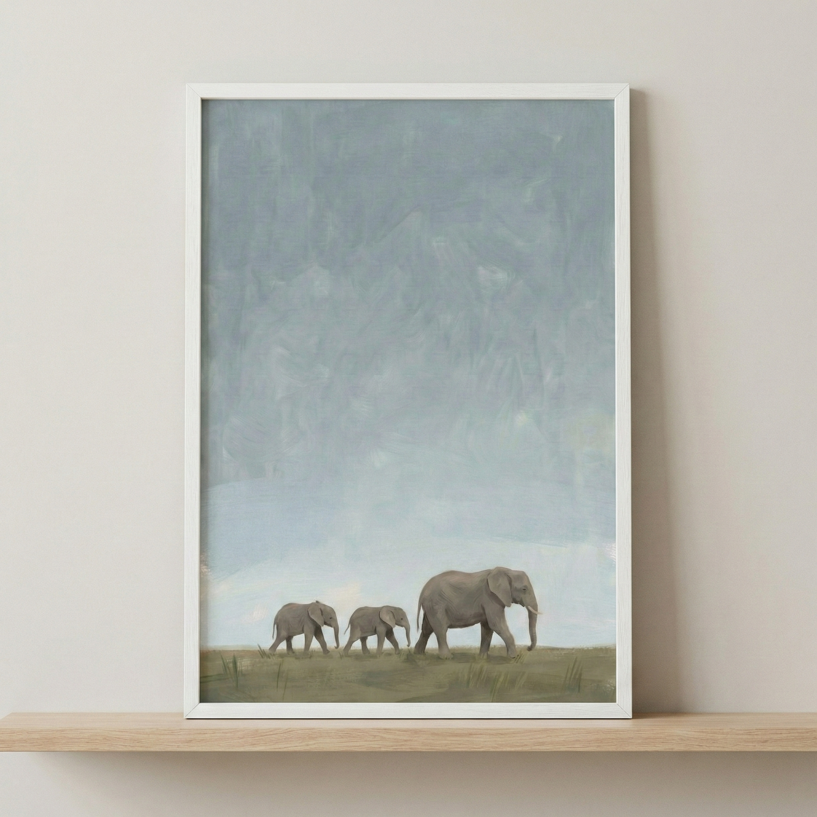 A piece of artwork in a thin white frame depicts three elephants walking in a single file across a flat, grassy horizon. A large elephant leads two smaller ones from right to left. The top two-thirds of the image is filled with a vast, textured sky in soft, muted shades of pale blue and grey. The overall style is minimalist and serene with subtle, visible textures. The framed image is displayed on a light wood shelf against a neutral-colored wall.