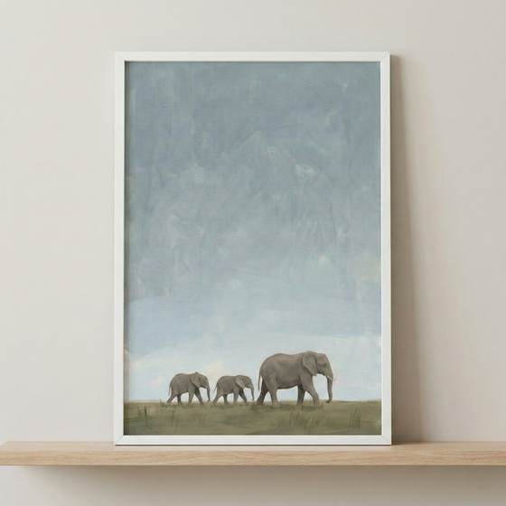 A piece of artwork in a thin white frame depicts three elephants walking in a single file across a flat, grassy horizon. A large elephant leads two smaller ones from right to left. The top two-thirds of the image is filled with a vast, textured sky in soft, muted shades of pale blue and grey. The overall style is minimalist and serene with subtle, visible textures. The framed image is displayed on a light wood shelf against a neutral-colored wall.