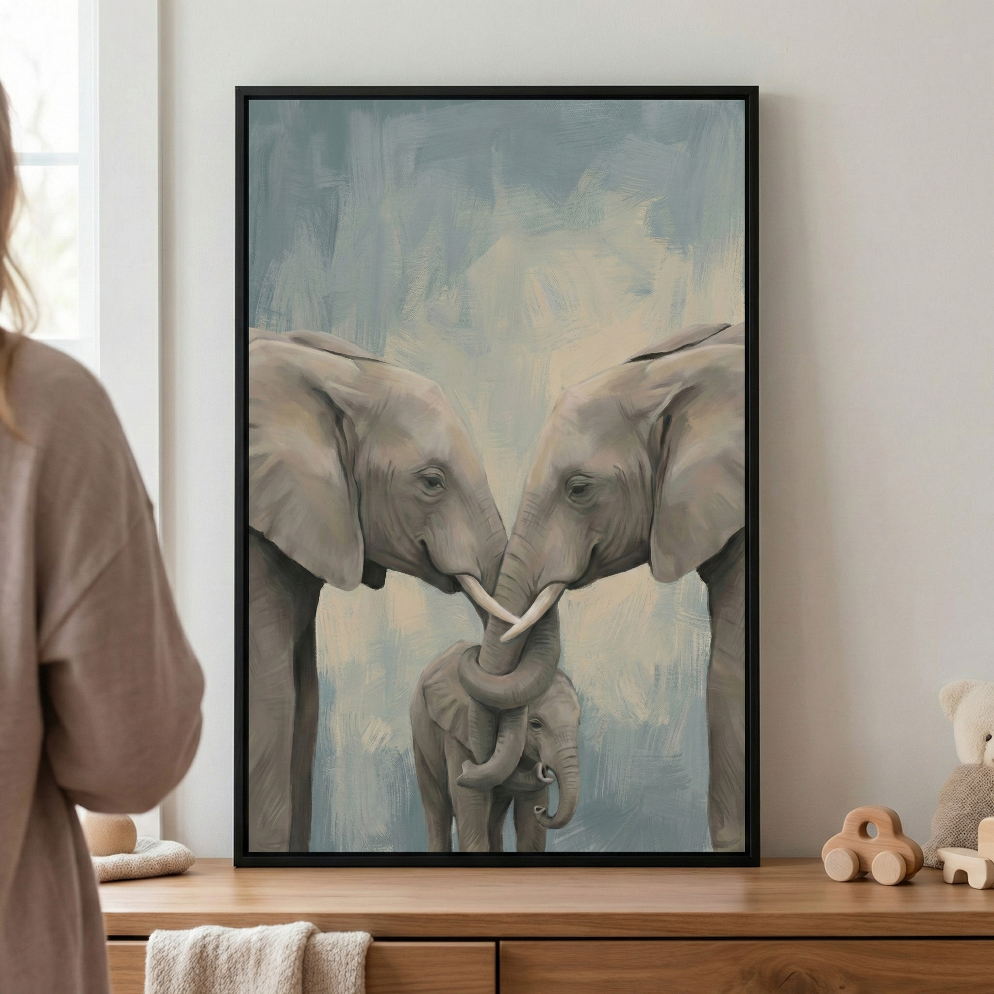 A vertically oriented, framed artwork hangs on a light-colored wall above a wooden chest of drawers. The piece depicts a family of three elephants: two large adults and a small calf. The adult elephants stand facing each other, their heads touching and their long trunks intertwined over the top of the calf's head in a protective gesture. The illustration features soft, visible textures and a muted color palette of grays, blues, and creams. The artwork is enclosed in a minimalist black frame.