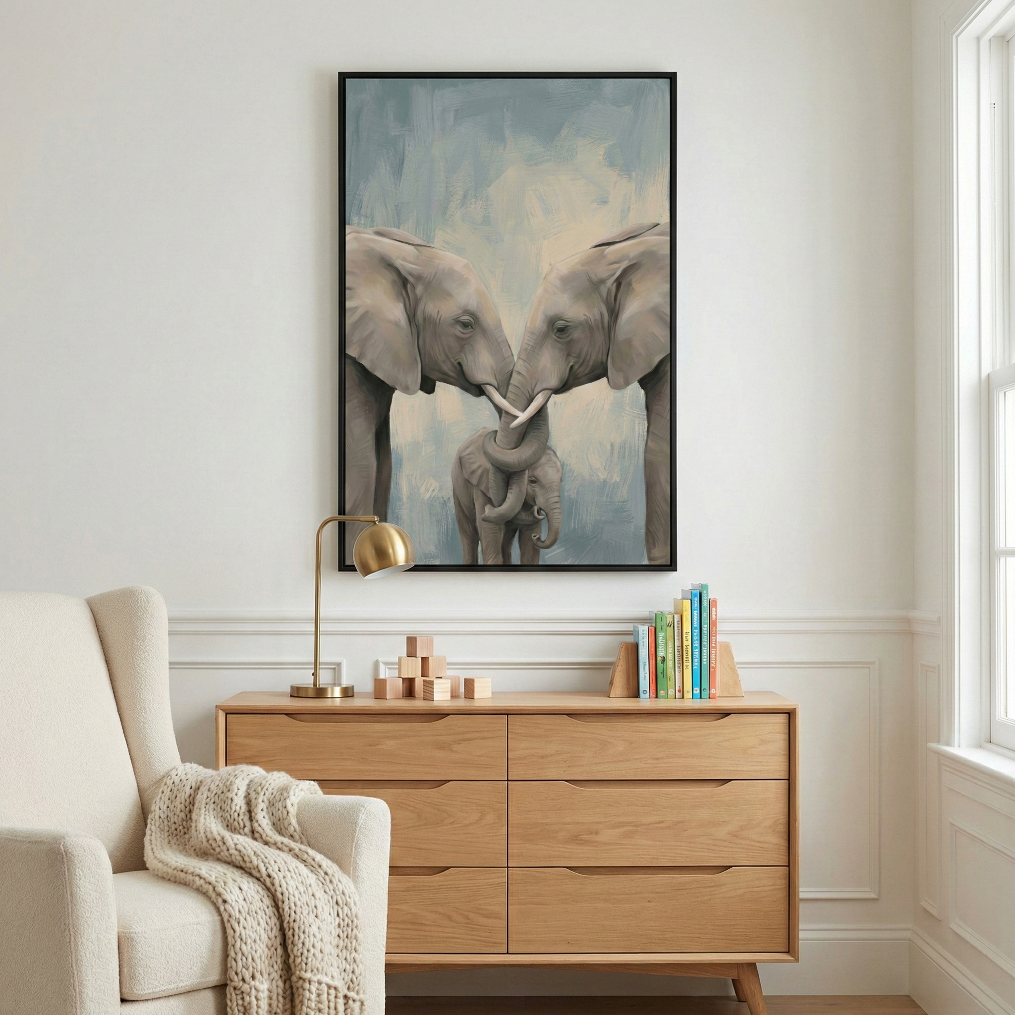 A vertical canvas artwork framed in black hangs on a white wall above a light wood dresser. The artwork shows two adult elephants facing each other, their trunks affectionately wrapped around a smaller baby elephant standing between them against a soft blue and beige background. On the dresser is a gold desk lamp, a stack of wooden blocks, and a small collection of colorful books. To the left is part of an off-white armchair with a textured cream throw blanket. The room is brightly lit.