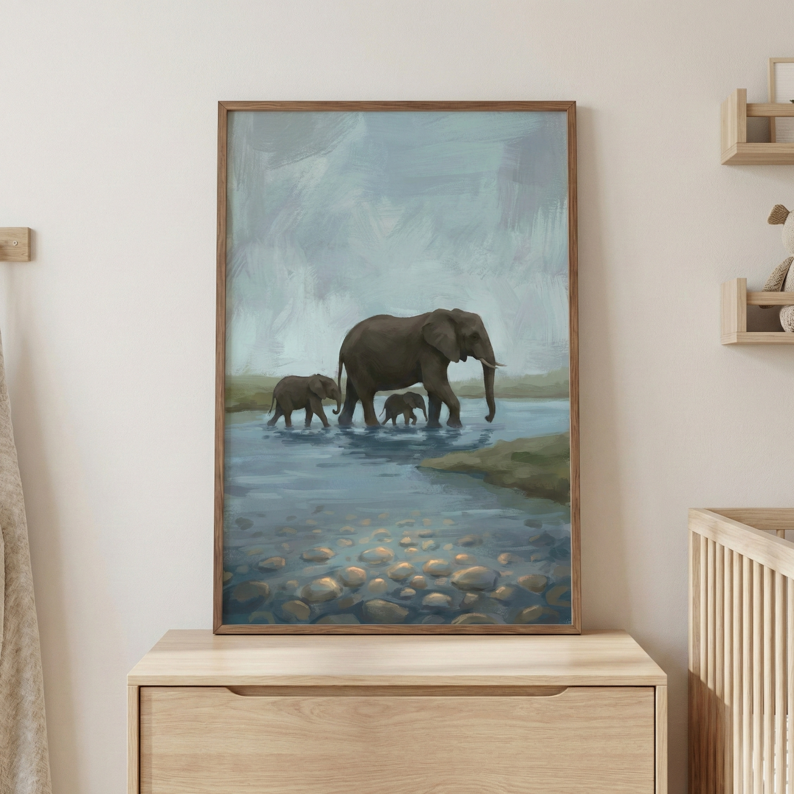 A vertical framed artwork sits on a light wood dresser, depicting a serene scene of three elephants wading through a shallow body of water. A large adult elephant leads the way, followed closely by two smaller calves. The water reflects a soft, overcast sky, and smooth river stones are visible through the clear ripples in the foreground. The piece is set within a thin wooden frame that matches the nearby furniture. 