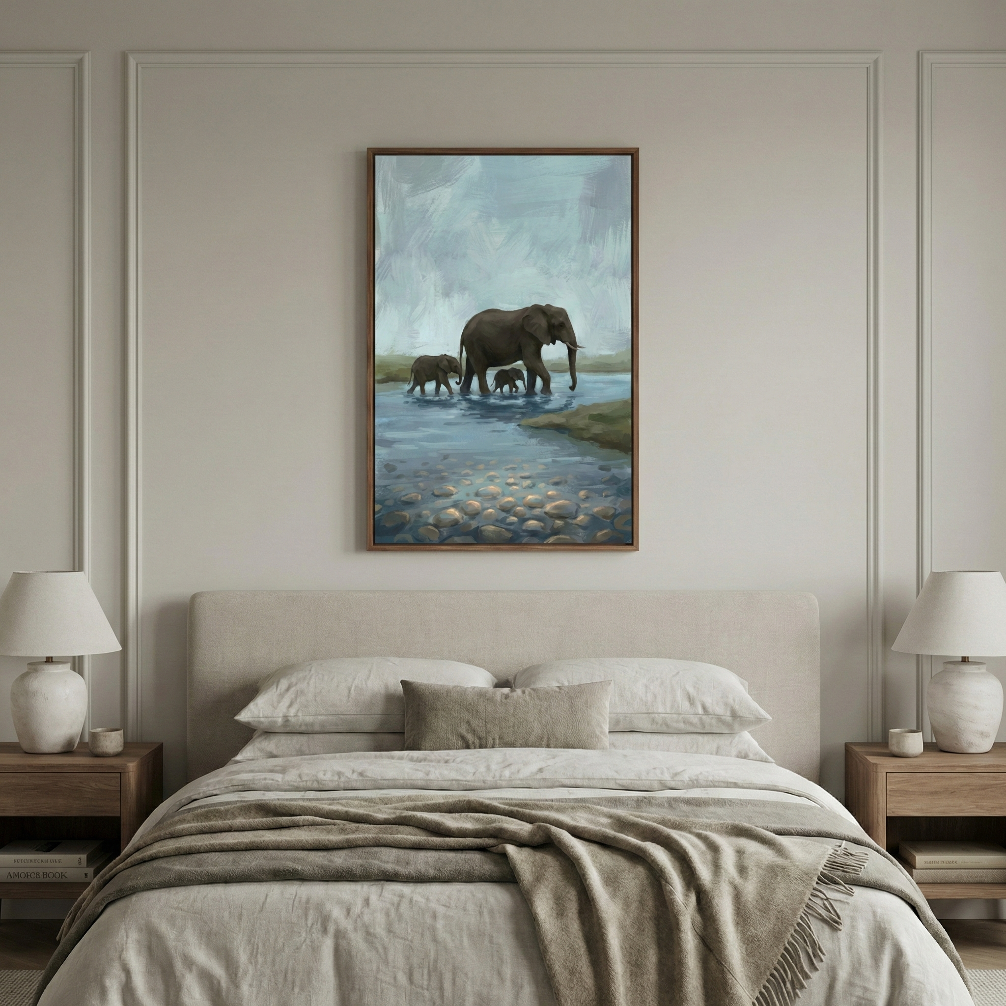 A serene bedroom setting showcases a neutral-toned bed with layered linens. On either side, matching wooden nightstands support elegant white lamps. Centered above the bed, a large framed art piece portrays a family of elephants—an adult and two calves—crossing a shallow, pebbled river under an atmospheric sky. Subtle wall molding adds a touch of architectural detail to the space, enhancing the peaceful and coordinated interior design.