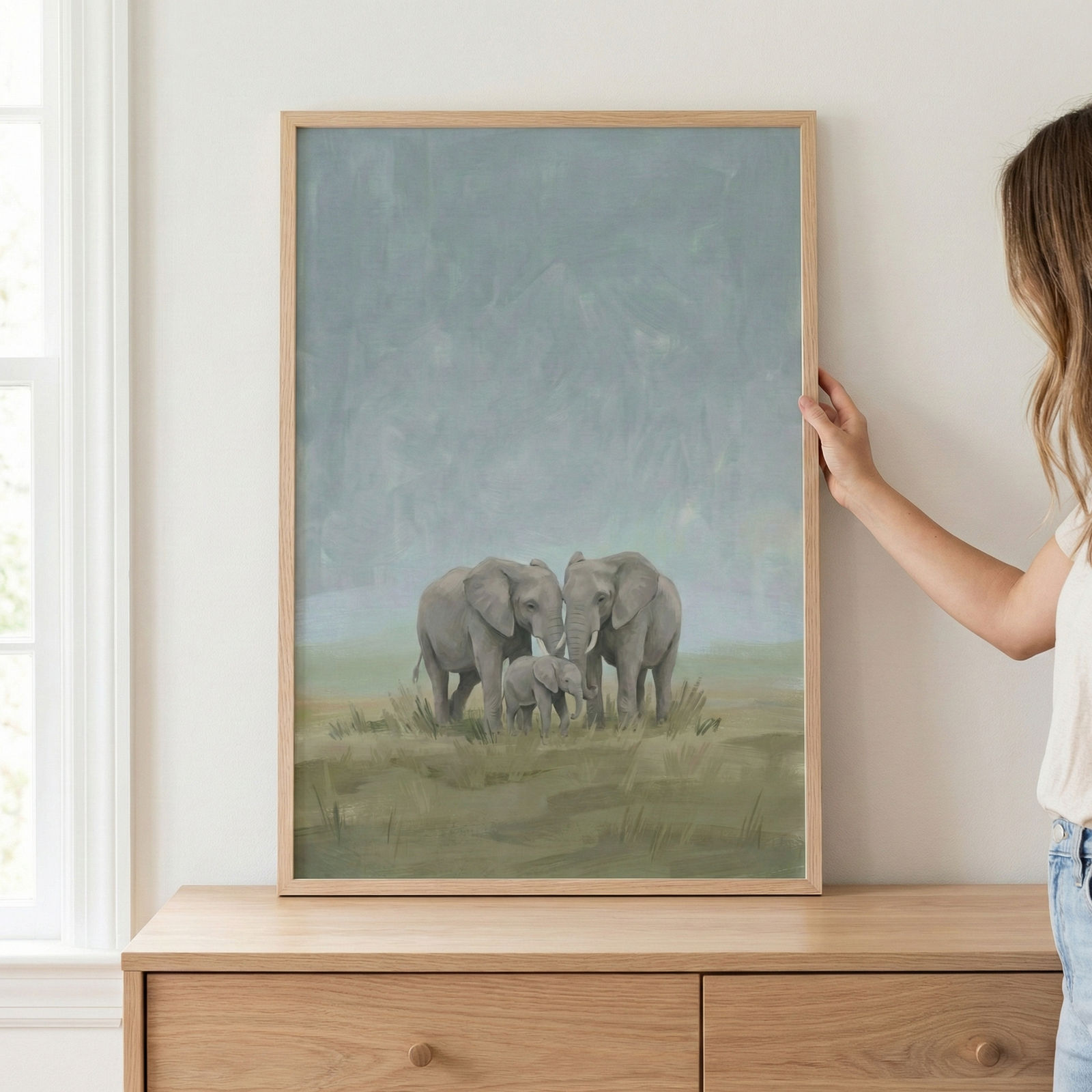 This vertical canvas art showcases a serene illustration of an elephant family. Two large adult elephants stand protectively over a small calf in the center. The figures are rendered with soft, textured strokes against a muted landscape featuring a grassy foreground and a vast, hazy blue-grey sky. The artwork is housed in a simple, light-colored wooden frame. A person’s hand is visible on the right edge, holding the frame against a neutral-toned wall. 