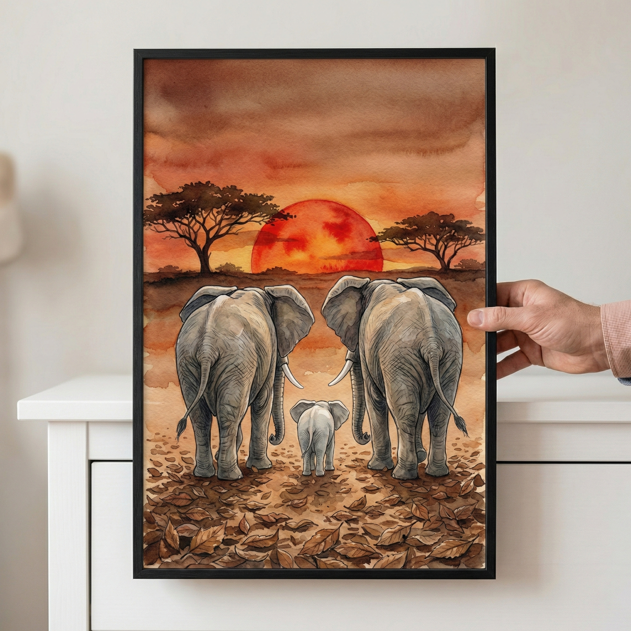 A framed watercolor-style illustration depicts an elephant family, consisting of two large adults and a small calf, walking toward a massive red sun on the horizon. The setting is a savanna with sparse, wide-canopied trees and a ground layered with fallen brown leaves. The sky is rendered in warm washes of orange, red, and brown. A person's hand is holding the black-framed artwork against a white backdrop.
