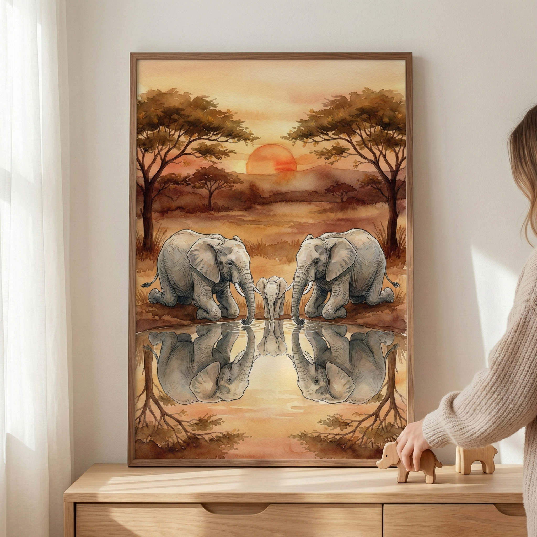 A framed watercolor-style illustration showing an elephant family at a watering hole during a warm sunset. Two adult elephants and a small calf are gathered at the water's edge, with their reflections clearly visible in the still water. The scene is set against a savanna landscape with a large, orange sun on the horizon and sparse trees. The frame rests on a light wood cabinet next to a person's hand holding a small wooden toy elephant.