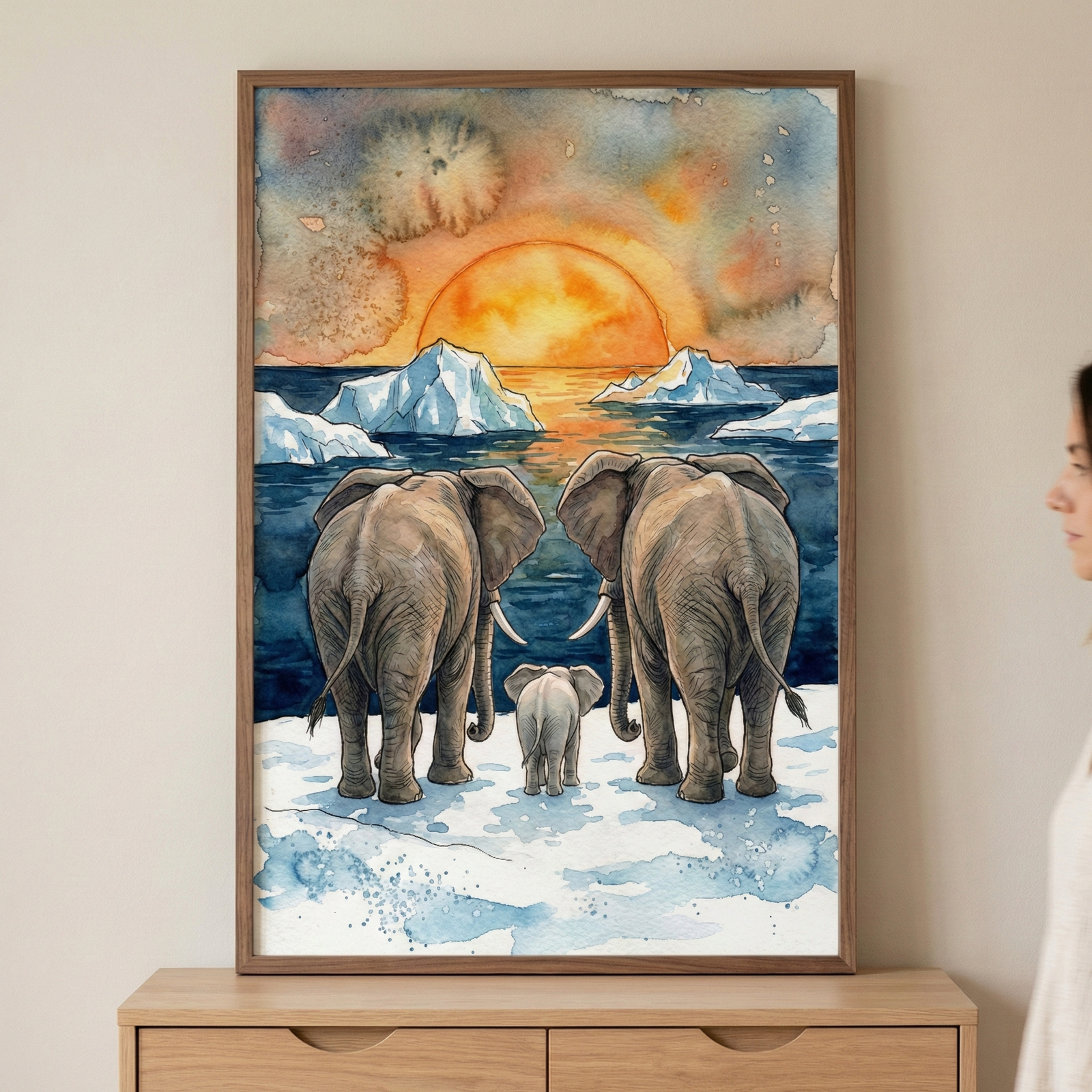 A framed watercolor illustration of two large elephants and a small calf standing on an icy landscape, looking out at a sunset over a sea with floating icebergs. The sky is filled with vibrant orange, yellow, and blue washes, centered around a large glowing sun. The artwork is held in a thin brown wooden frame and is displayed on a light wall above a wooden dresser, with a person’s profile partially visible on the right.
