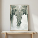 A vertically oriented, light-wood framed artwork featuring a soft-toned illustration of two large adult elephants facing each other with their trunks delicately intertwined. Tucked between them at the bottom is a small baby elephant. The illustration uses muted sage green and gray hues with thin line work against a white background that transitions into a misty, cloud-like base. The frame rests on a beige cushioned wooden bench positioned against a neutral, light-colored wall.