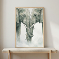 A vertically oriented, light-wood framed artwork featuring a soft-toned illustration of two large adult elephants facing each other with their trunks delicately intertwined. Tucked between them at the bottom is a small baby elephant. The illustration uses muted sage green and gray hues with thin line work against a white background that transitions into a misty, cloud-like base. The frame rests on a beige cushioned wooden bench positioned against a neutral, light-colored wall.