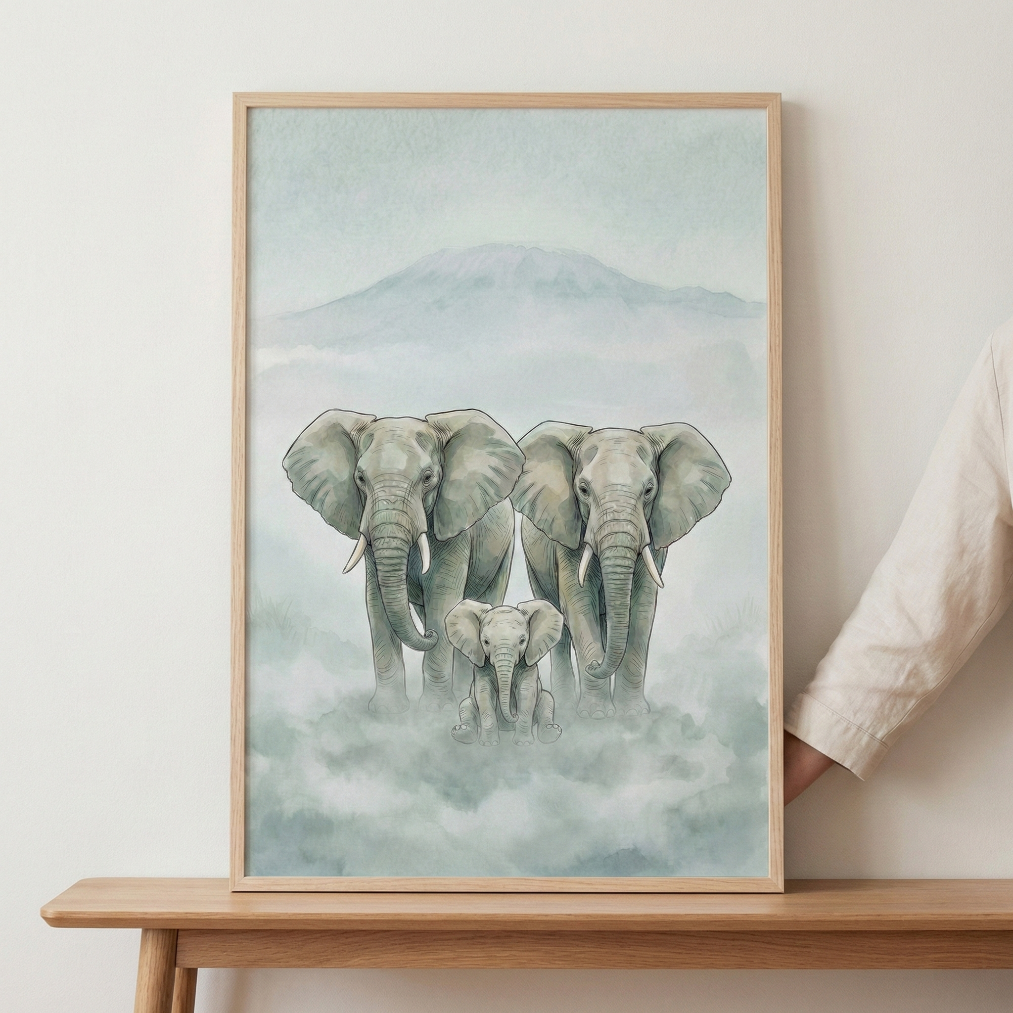 A framed illustration of three elephants—two adults and a calf—set against a misty, mountainous landscape. The art uses soft, muted colors and is displayed in a light wood frame resting on a wooden table. A hand is visible holding the side of the frame.