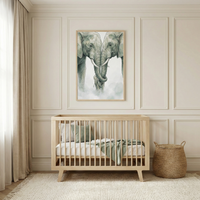 A serene nursery scene featuring a light wood crib with sage green and white bedding. Above the crib hangs a large framed watercolor-style illustration of two adult elephants with their trunks intertwined over a baby elephant. To the right of the crib is a round woven storage basket. The room is decorated in soft neutral tones with elegant wall molding, long beige curtains, and a textured cream rug.