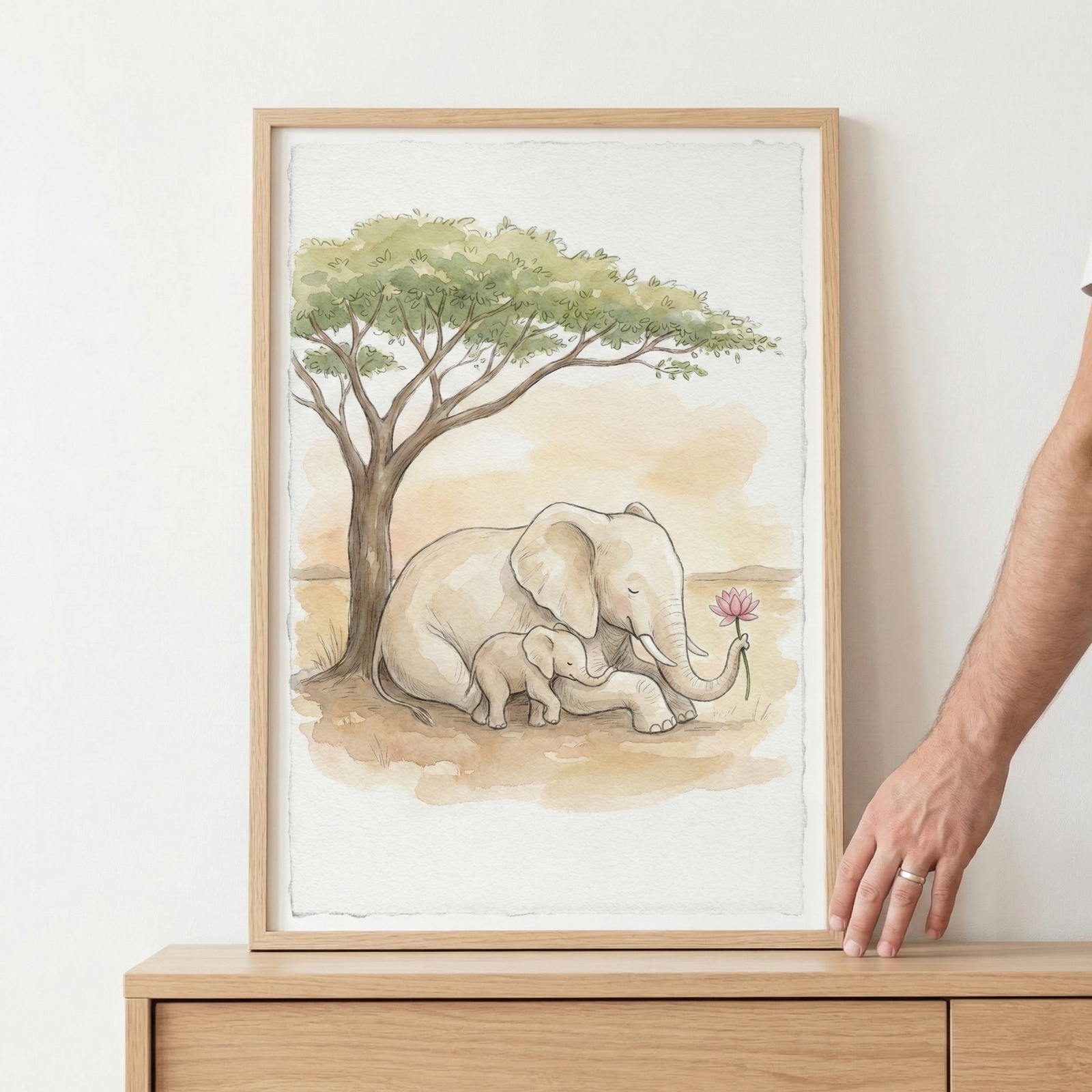 A framed watercolor-style illustration featuring an elephant and its calf nestled together. They are resting on the ground beneath the sprawling green canopy of a savanna tree. The larger elephant gently holds a single pink flower in its trunk, while the small calf sits close to its side. The artwork uses a soft palette of earthy browns, tans, and greens. The piece is housed in a light-colored wooden frame, positioned on a wooden surface against a plain white wall.