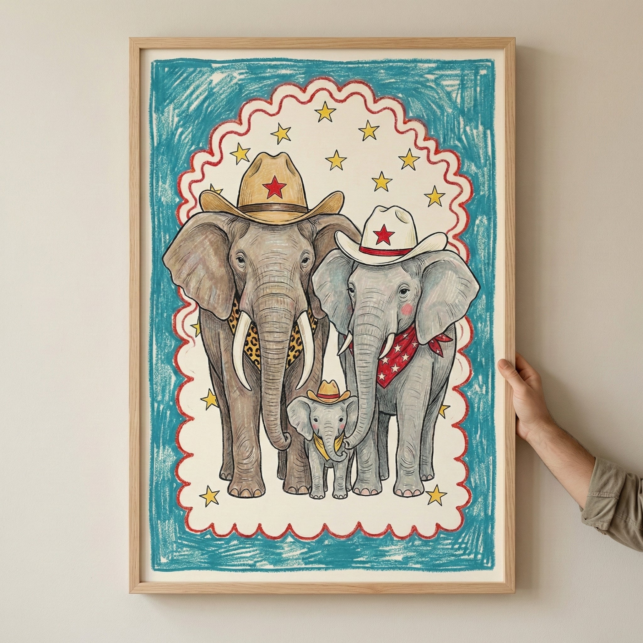 Framed illustration of three elephants dressed in western cowboy attire, including hats with red stars, a leopard print scarf, and a red bandana, with a colorful border of stars and blue scribble background. A hand is holding the frame against a neutral wall.