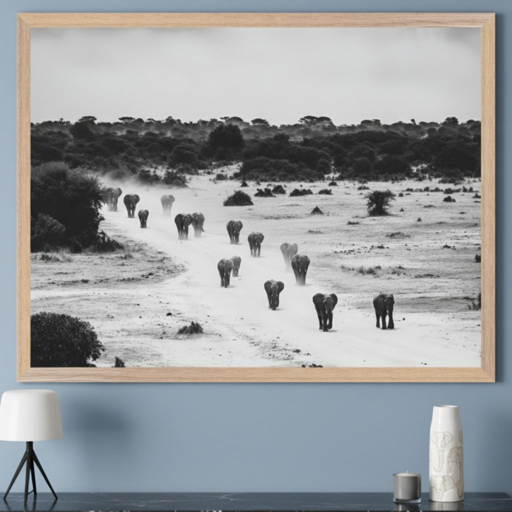 Modern living room with a large light wood framed black and white photo of elephants walking on a dirt road through a savanna, centered above a black marble console table. A white lamp and a white vase with an elephant pattern are on the console.