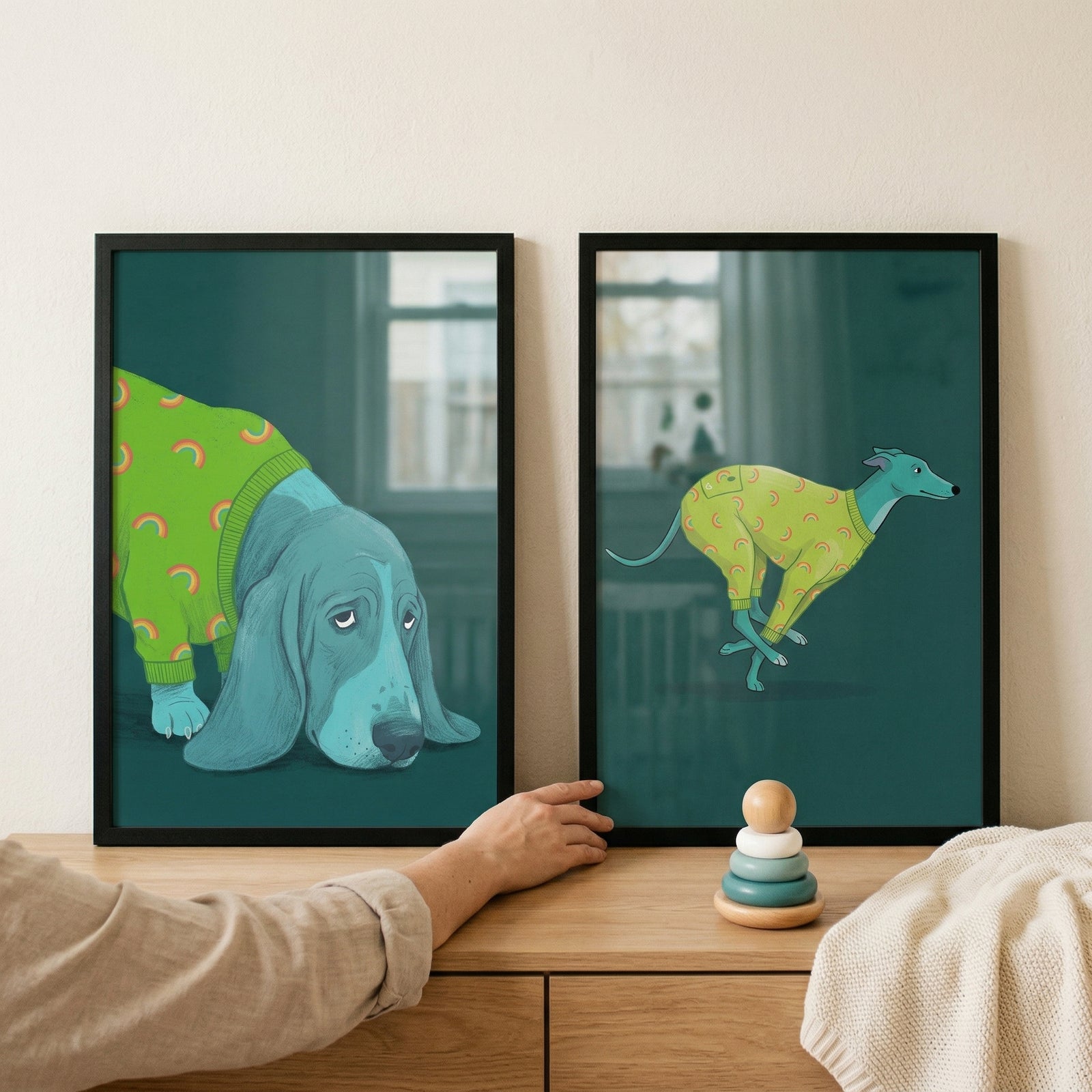 Two black-framed illustrations rest on a light wood dresser against a plain wall. The left artwork features a large, light-blue cartoon dog with long, drooping ears and a somber expression, wearing a bright green sweater patterned with small orange rainbows. 