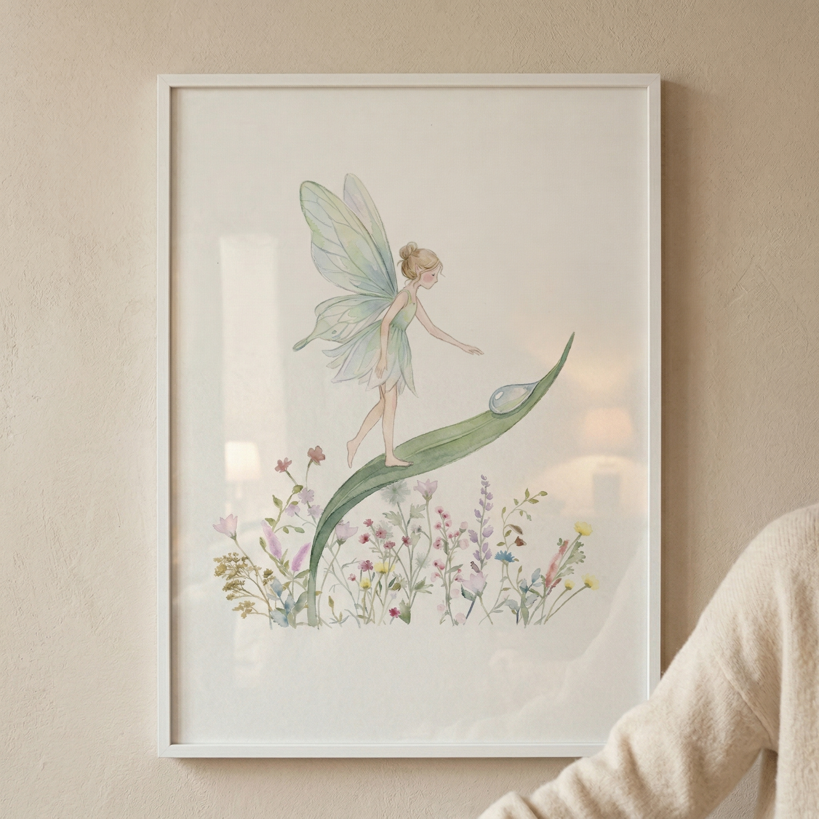 A framed watercolor illustration of a whimsical fairy with translucent wings and a light green dress walking along a large, curved leaf. She reaches toward a single dewdrop on the leaf's surface. Below the leaf is a collection of colorful wildflowers in muted tones. The artwork is housed in a simple white frame and hung on a textured beige wall, with a person in a soft-knit sweater partially visible on the right.