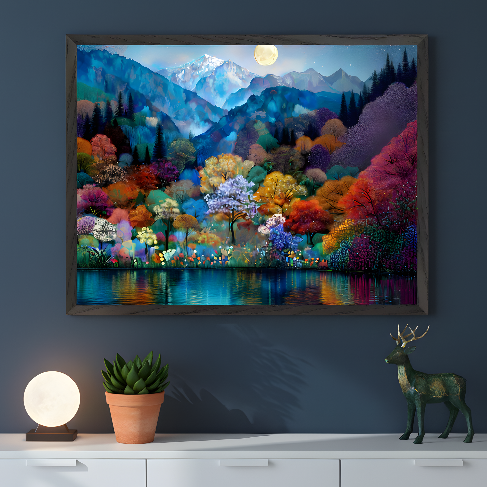 Framed wall art of a vibrant forest scene hangs on a dark blue wall, showing colorful trees reflected in a calm lake beneath a glowing full moon and snowy mountains. Below the artwork, a white console holds a small potted plant, a round glowing lamp, and a green deer figurine, creating a serene and artistic display.