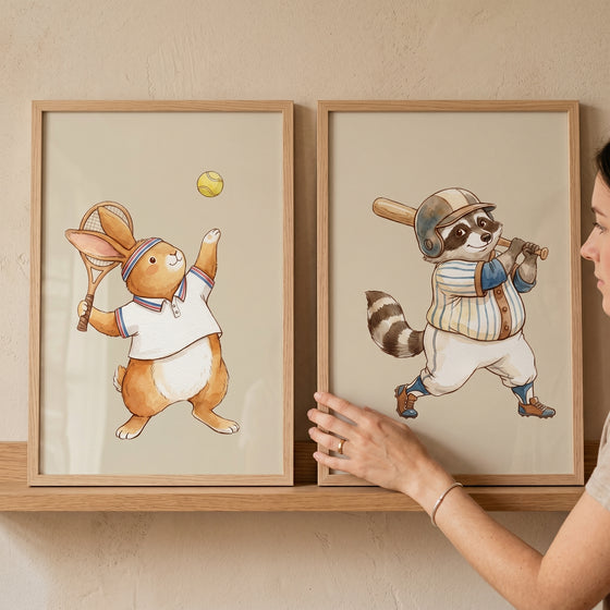 Energetic watercolor athletic animals wall art set of 2 shows a rabbit playing tennis and a raccoon playing baseball.