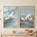 Two framed pieces of art are hung side-by-side on a light-colored wall above a wooden dresser. On the dresser's surface, there are folded neutral-colored cloths, some small items, and a wicker basket containing a wooden bear rattle and blocks. A hand is visible resting on the right side of the dresser.
The artwork on the left depicts three white dogs with a distinctive, curved head profile—one larger dog and two smaller ones—standing on a green, grassy slope. 